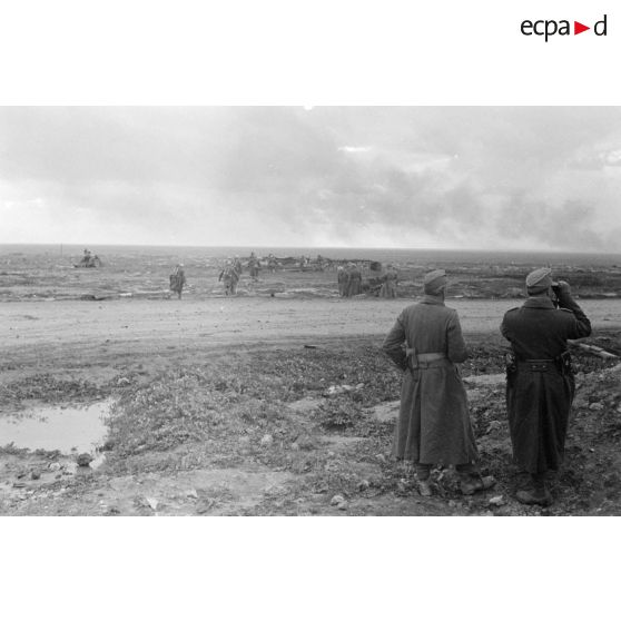 Deux officiers observent dans la direction des combats d'où se dégage un large nuage de fumée.