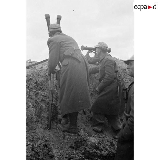 Protégés derrière un parapet ou dans une tranchée, des officiers et un soldat observent les combats avec un binoculaire et un télémètre.