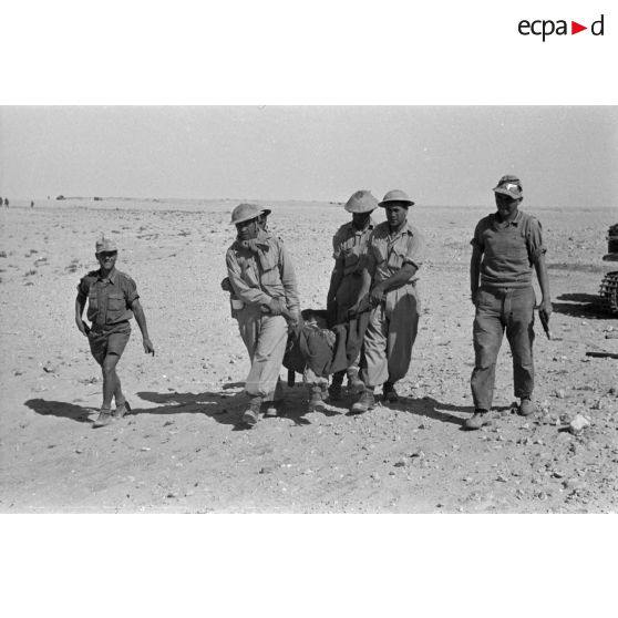 Des prisonniers transportent un blessé installé dans une couverture, gardés par deux soldats allemands armés de pistolets Walther-P-38.