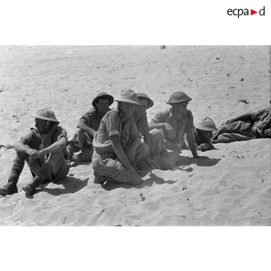 Des prisonniers britanniques accroupis et allongés dans le sable.