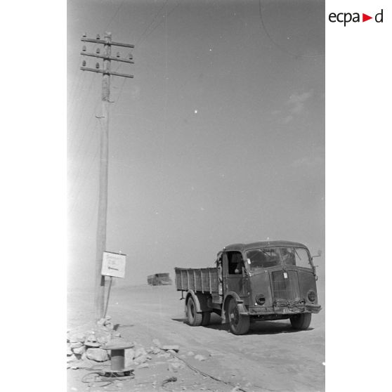 Passage de camions le long de poteaux télégraphiques et à coté d'un panneau indiquant un point de rassemblement.