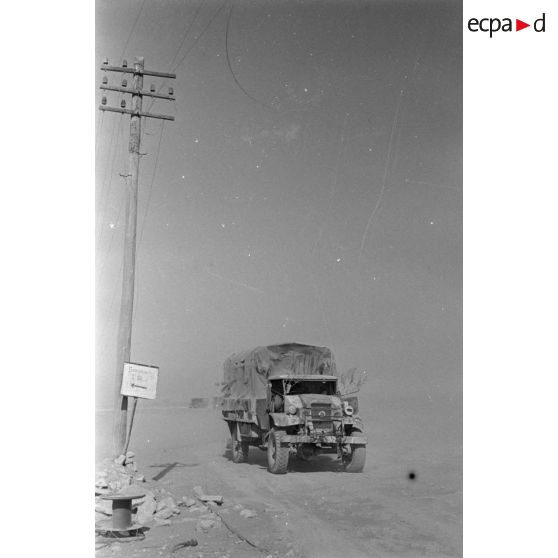 Passage de camions le long de poteaux télégraphiques et à coté d'un panneau indiquant un point de rassemblement.