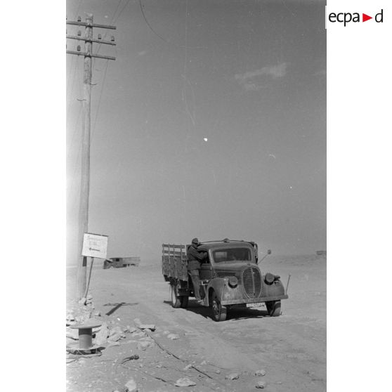 Passage de camions le long de poteaux télégraphiques et à coté d'un panneau indiquant un point de rassemblement.