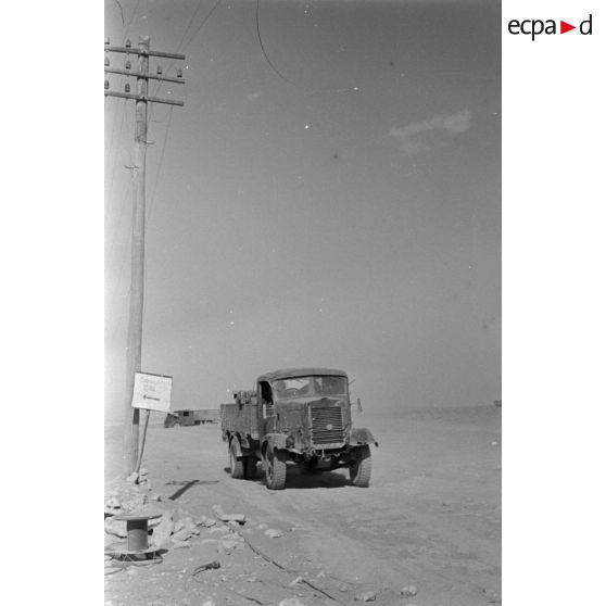 Passage de camions le long de poteaux télégraphiques et à coté d'un panneau indiquant un point de rassemblement.