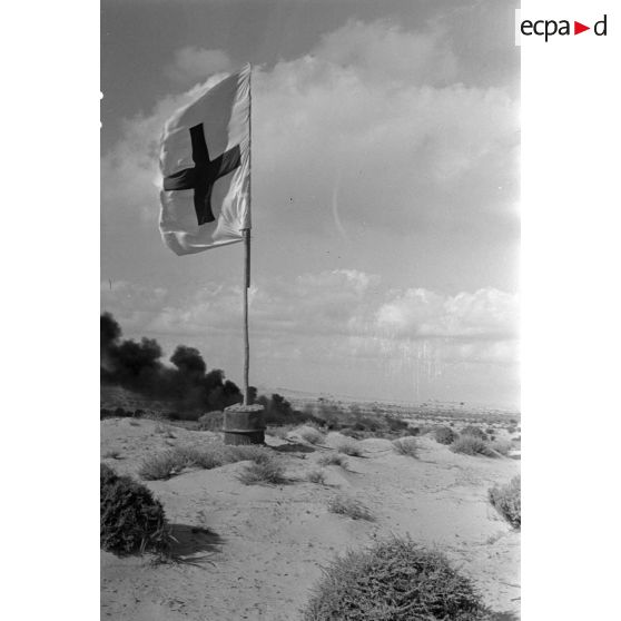 Des débris d'un avion en feu près d'un drapeau de la croix rouge dans le désert.