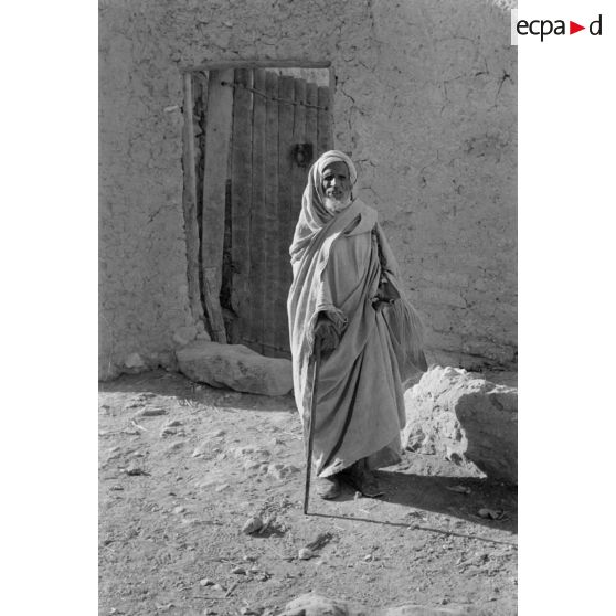 Portrait en pied d'un vieil homme devant l'entrée d'une habitation.