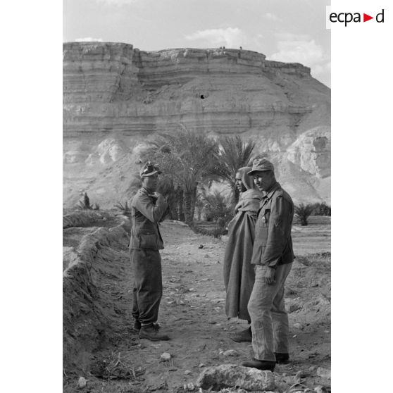 Un lieutenant (Oberleutnant) et un caporal (Gefreiter) discutent avec des habitants du village au milieu des cultures de l'oasis.