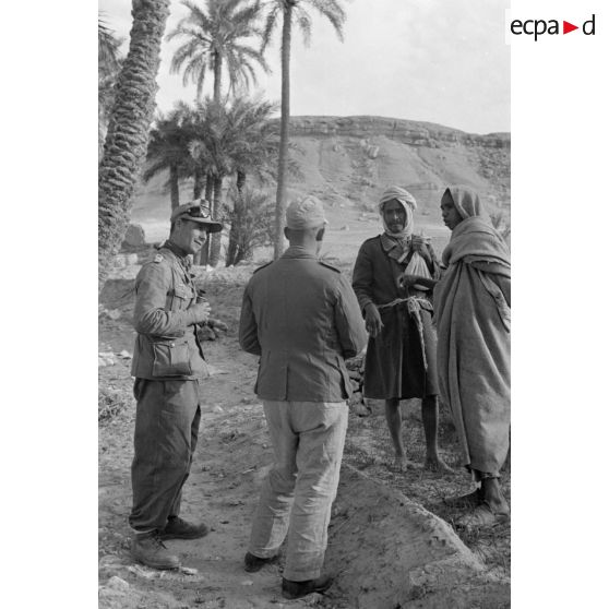 Un lieutenant (Oberleutnant) et un caporal (Gefreiter) discutent avec des habitants du village au milieu des cultures de l'oasis.