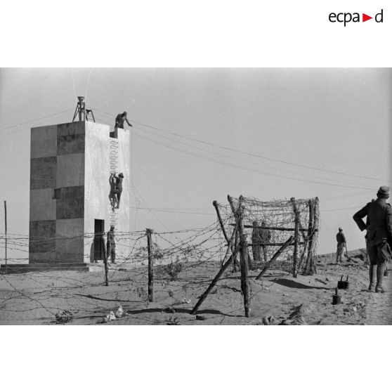 Une tour entourée de barbelés et des soldats se préparent à poser des mines.