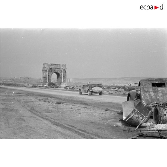 Une voiture Kübelwagen progressant le long de la route, on reconnaît l'Arc de Dioclétien près d'une épave de camion et des ruines romaine de Sufetula.