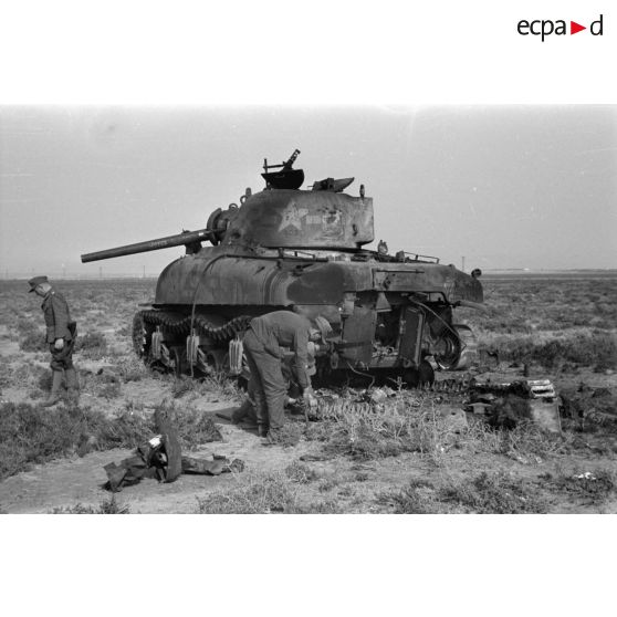 Des soldats s'affairent autour de la carcasse d'un char américain Sherman M4A1 détruit, sur le canon de 75 mm, on note le prénom Joyce.