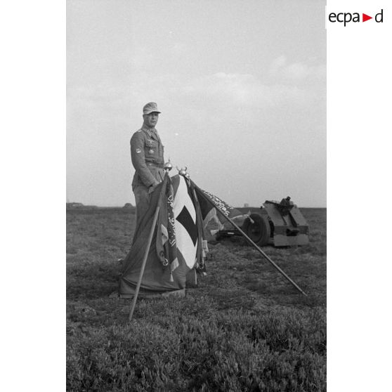 Un officier allemand fait une allocution depuis une tribune improvisée composée d'un drapeau à croix gammée et des fanions des unités.