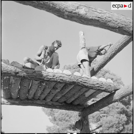 Au moyen d'un marteau, un soldat de la 3/131e régiment d'infanterie (RI) cloue "le plancher" constitué de rondins de bois taillés d'une structure en bois en cours de construction.