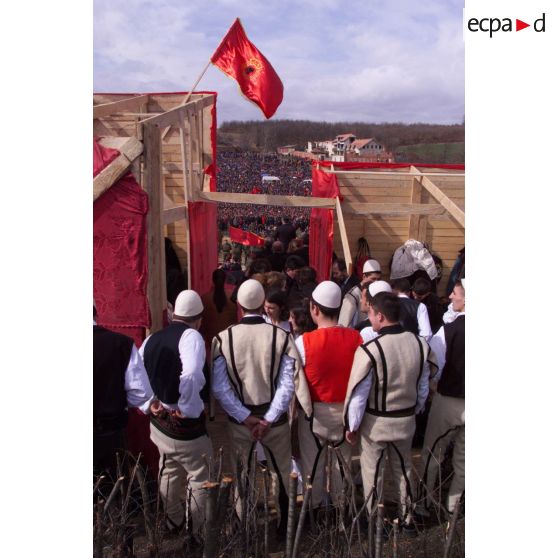 Vue des coulisses avec un groupe folklorique de dos sous un drapeau de l'armée de libération du Kosovo (UCK).