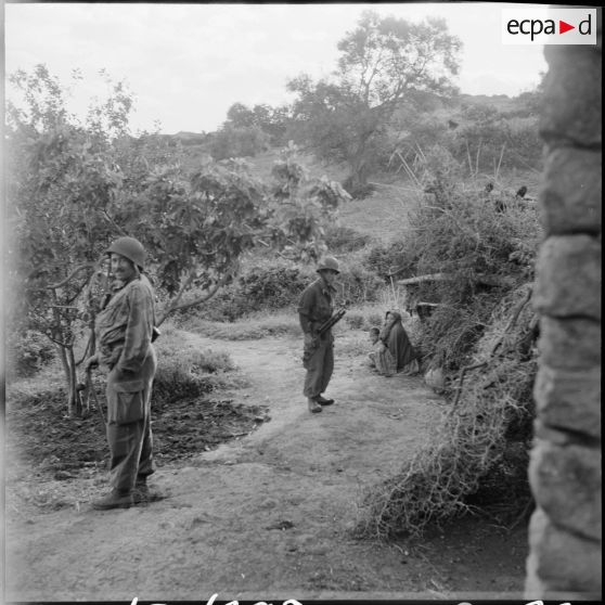 Deux soldats surveillent la rue d'un douar en arrière plan, une femme assise et un enfant.