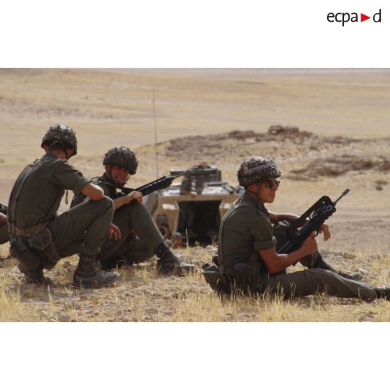 Trois soldats portant le casque lourd et armés de fusil d'assaut FAMAS sont assis au sommet d'une colline dans le désert lors d'un exercice de tir.