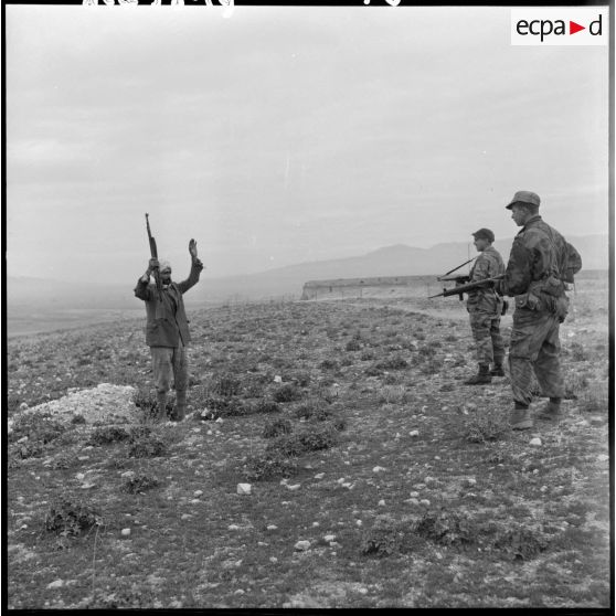 Un homme, mains en l’air et tenant un fusil, se rend devant deux soldats armés lors d’une opération héliportée sur le djebel Tarf.