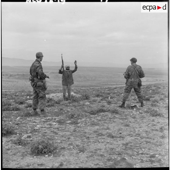 Un homme, mains en l’air et tenant un fusil, se rend devant deux soldats armés lors d’une opération héliportée sur le djebel Tarf.