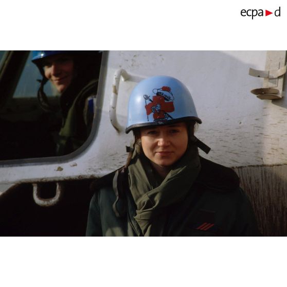 Portrait d'une infirmière du Service de Santé des Armées posant devant un véhicule de l'avant blindé (VAB) et arborant un casque bleu.