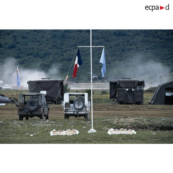 Mât des couleurs avec drapeau français et ONU, Peugeot P4 au parking, douches de campagne et atterrissage d'un hélicoptère Puma SA 330 sur le camp de Lipa.