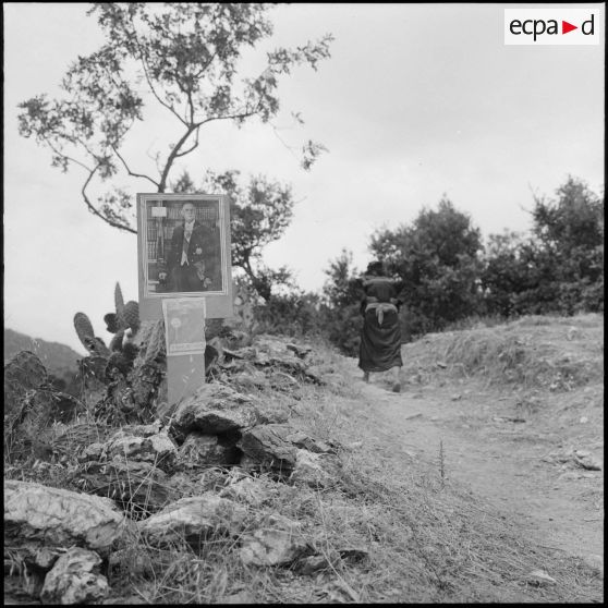 Une femme passe sur une route de haute montagne au bord de laquelle est placée une photographie du général de Gaulle.