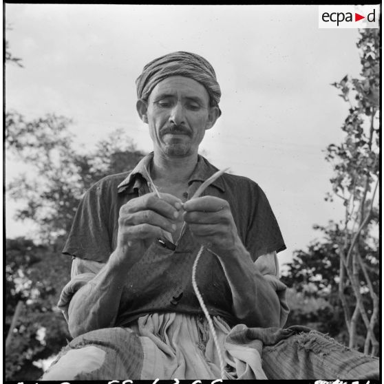 Portrait d'un homme qui confectionne des cordelettes avec des feuilles sèches.