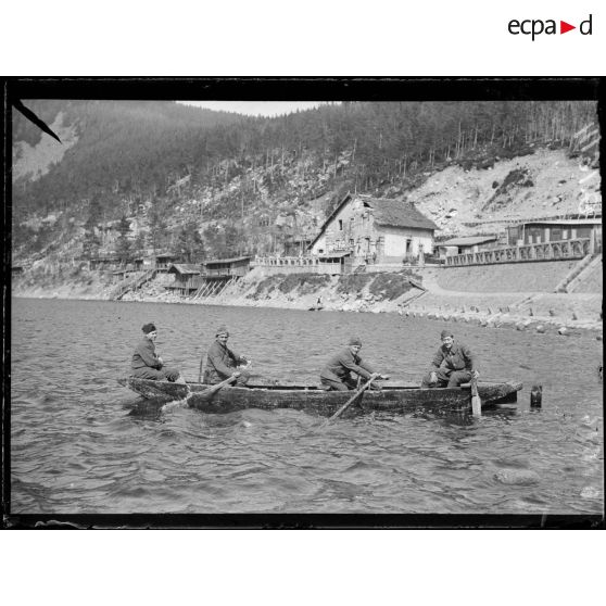 [Quatre soldats sont à bord d'une embarcation au pied d'une montagne, où se trouvent des habitations.]