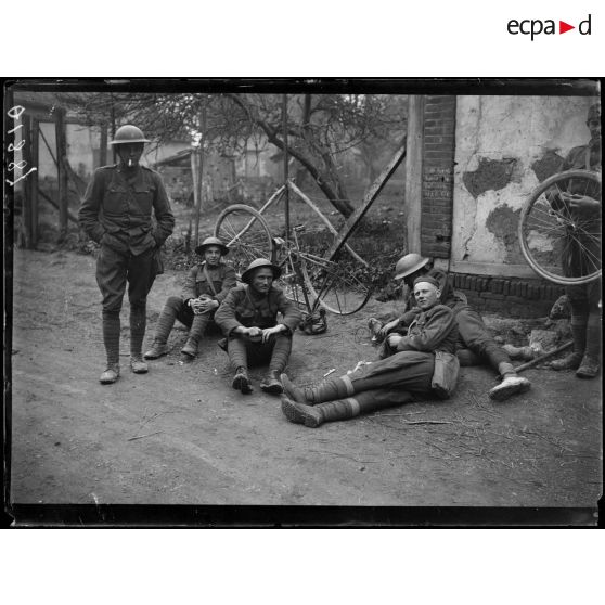 [Des soldats sont assis et allongés sur le bord d'une route de village, pendant que l'un d'entre eux répare la roue d'un vélo.]