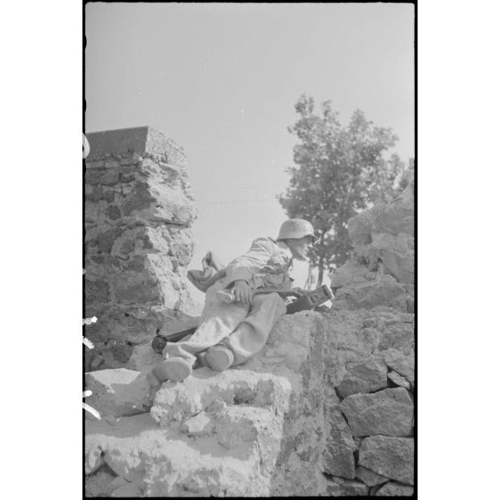 Dans le secteur de Catane, chargé d'une caisse de munitions pour une mitrailleuse, un fantassin du Panzer-Grenadier-Regiment Hermann Goering observe le front à travers un mur effondré.