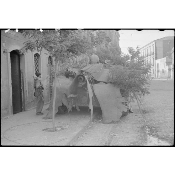 Garé ou embusqué sur un trottoir, le long des habitations, un Sturmgeschütz III de la III./Panzer-Regiment Hermann Goering est recouvert d'une tôle ondulée ou caoutchoutée, d'une paillasse et de branches.