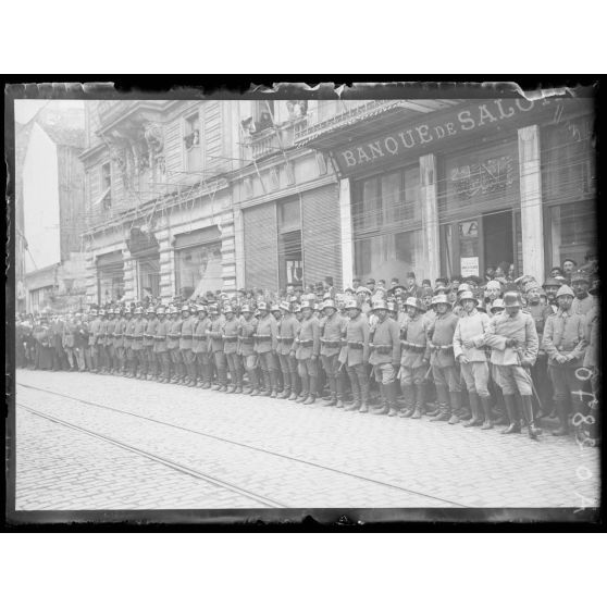 [Population civile et troupes militaires attendent le passage d'un convoi, en centre ville.]