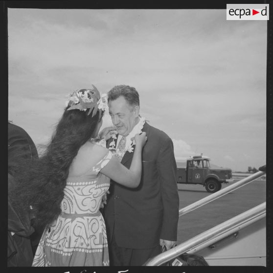 Le capitaine de vaisseau Bernoud, commandant du site de Moruroa (Mururoa), arrivant à Faa'a, l'aéroport de Papeete, se voit offrir un collier de fleurs par une hôtesse polynésienne en tenue traditionnelle.