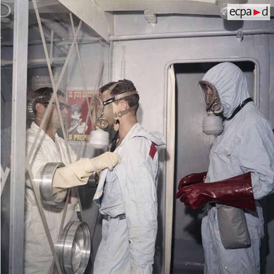 Protégé par un vitrage, un membre de l'équipe de décontamination ôte la tenue de protection d'un personnel à bord du bâtiment de soutien santé (BSS) Rance (A618), spécialisé dans la sécurité radiologique.