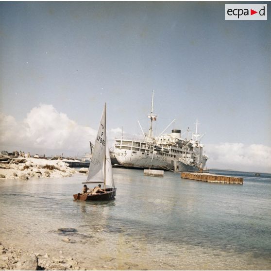 Au port de Moruroa, deux personnels naviguent près du rivage en dériveur, non loin du bâtiment-base Maurienne (A637) et du bâtiment-citerne à eau Liamone ex-Arrosoir (A750) à quai.