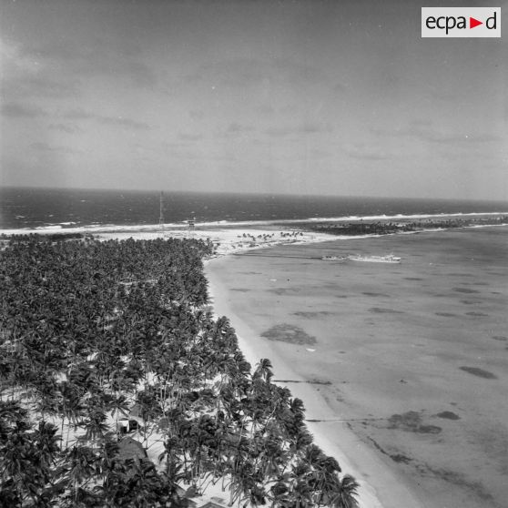 Vue aérienne du site du poste de commande de tir (PCT) Anémone sur l'atoll de Moruroa. Au premier plan une zone vie avec des fare de Moruroa. Au bout de la jetée, un bâtiment-atelier (peut-être L9082).