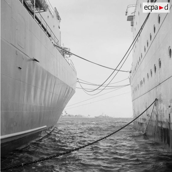 Vue entre les coques de deux bâtiments (peut-être le bâtiment-base (BB) Médoc (A612)) à quai au port de Moruroa de la force Alpha au mouillage dans le lagon.