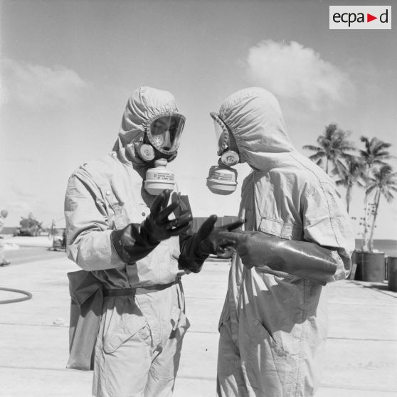 Sur une aire de lavage du service de décontamination de Hao, discussion entre deux personnels en tenue de protection après un tir d'essai nucléaire, à proximité de barils. Les masques portent l’inscription «protection assurée contre les poussières et l’iode radioactif , licence CEA».