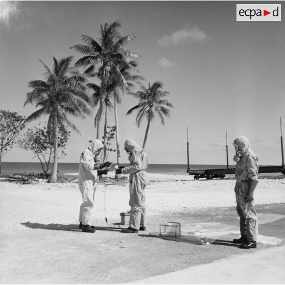 Sur une aire du service de décontamination de Hao, des personnels en tenue de protection testent la radioactivité de matériel avec un détecteur après un tir d'essai nucléaire.