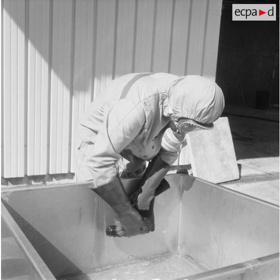 Devant un hangar du service de décontamination de Hao, un personnel en tenue de protection brosse et lave une botte dans un bac après un tir d'essai nucléaire.