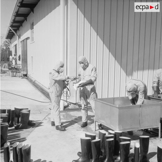 Devant un hangar du service de décontamination de Hao, des personnels en tenue de protection lavent des bottes dans un bac et contrôlent leur radioactivité avec un détecteur après un tir d'essai nucléaire.