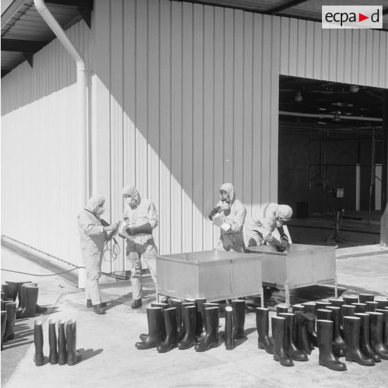 Devant un hangar du service de décontamination de Hao, des personnels en tenue de protection lavent des bottes dans un bac et contrôlent leur radioactivité avec un détecteur après un tir d'essai nucléaire.