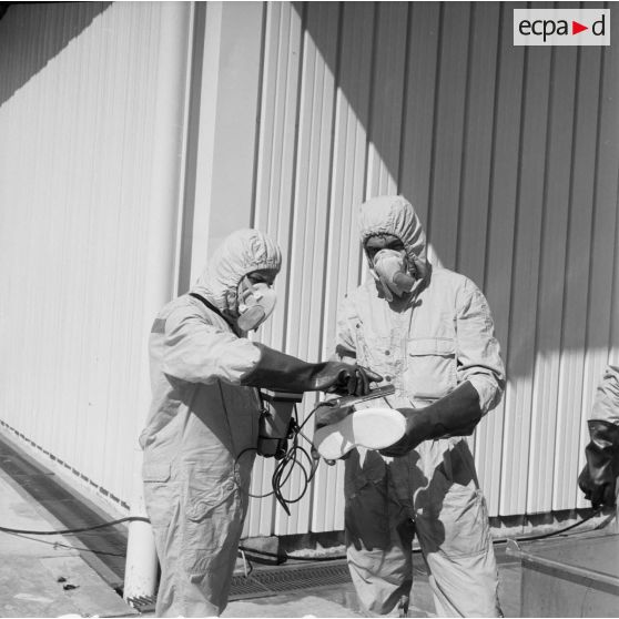 Devant un hangar du service de décontamination de Hao, des personnels en tenue de protection contrôlent la radioactivité sur les bottes lavées avec un détecteur après un tir d'essai nucléaire.