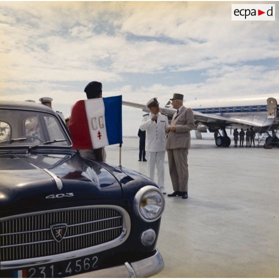 Accueil du général Charles De Gaulle, président de la République, par le vice-amiral Jean Lorain, commandement du groupe opérationnel des expérimentations nucléaires (GOEN), à l'aéroport de Moruroa. Au premier plan, la voiture officielle Peugeot 403 et le drapeau aux initiales du président.