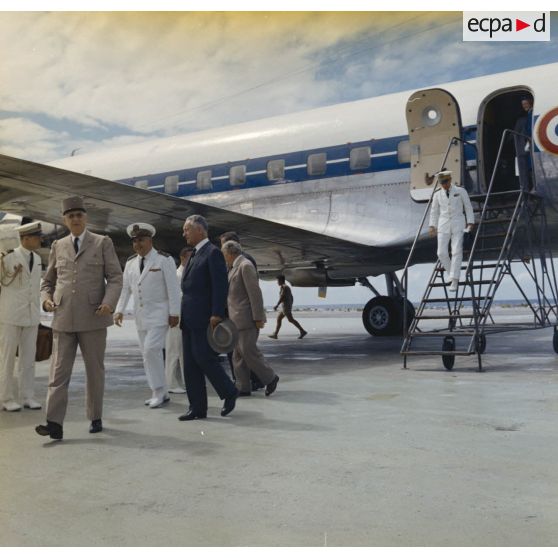 Le général Charles De Gaulle, président de la République, suivi de son aide de camp et de Pierre Messmer, ministre des Armées, au pied de l'avion à l'aéroport de Moruroa, accueilli par Jean Viard, directeur des essais, et le vice-amiral d’escadre Jean Lorain, commandant du groupe opérationnel des expérimentations nucléaires (GOEN).