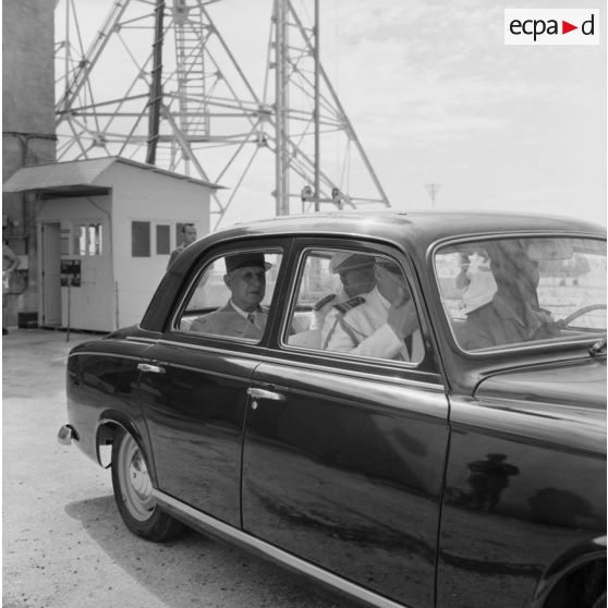 Au pied du bunker du poste d'enregistrement avancé (PEA) Denise, le général Charles De Gaulle, président de la République à bord de sa voiture Peugeot 403, aux côtés du vice-amiral Jean Lorain, commandant du groupe opérationnel des expérimentations nucléaires (GOEN), et de son aide de camp.