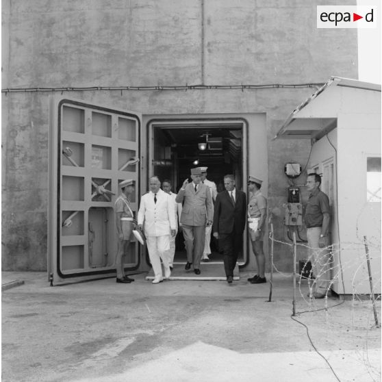 Le général Charles De Gaulle, président de la République, sort du bunker du poste d'enregistrement avancé (PEA) Denise, en compagnie du vice-amiral Jean Lorain, commandant du groupe opérationnel des expérimentations nucléaires (GOEN), et de Jean Viard, directeur des essais.