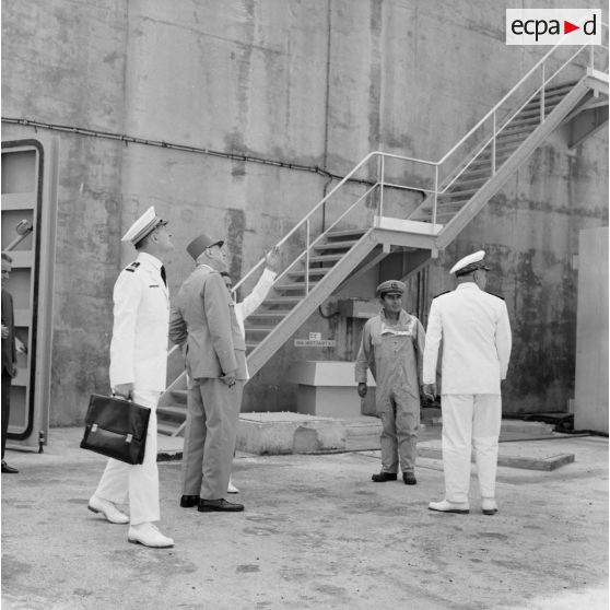 Le général Charles De Gaulle, président de la République, sort du bunker du poste d'enregistrement avancé (PEA) Denise, en compagnie du vice-amiral Jean Lorain, commandant du groupe opérationnel des expérimentations nucléaires (GOEN), et de Jean Viard, directeur des essais, suivi par son aide de camp.