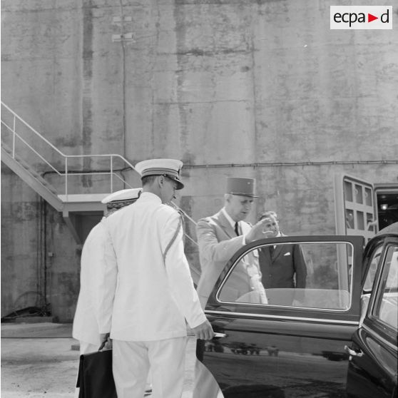 Au pied du bunker du poste d'enregistrement avancé (PEA) Denise, le général Charles De Gaulle, président de la République remonte en voiture, en présence du vice-amiral Jean Lorain, commandant du groupe opérationnel des expérimentations nucléaires (GOEN), d'Alain Peyrefitte, ministre de la Recherche scientifique et des Questions atomiques et spatiales, et de son aide de camp.