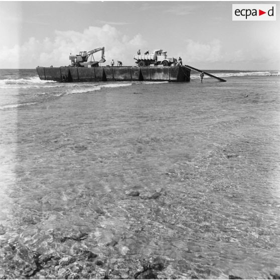 Ouvriers au travail pour le déchargement d'un engin pelleteuse et d'un camion benne d'un bâtiment sur le rivage. Construction du site de Moruroa.