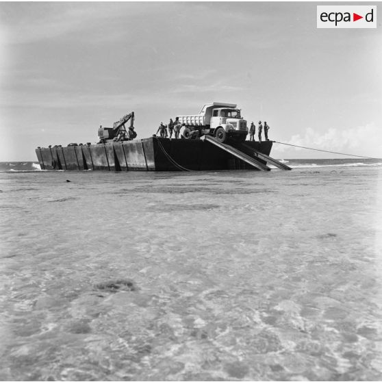 Ouvriers au travail pour le déchargement d'un engin pelleteuse et d'un camion benne d'un bâtiment sur le rivage. Construction du site de Moruroa.